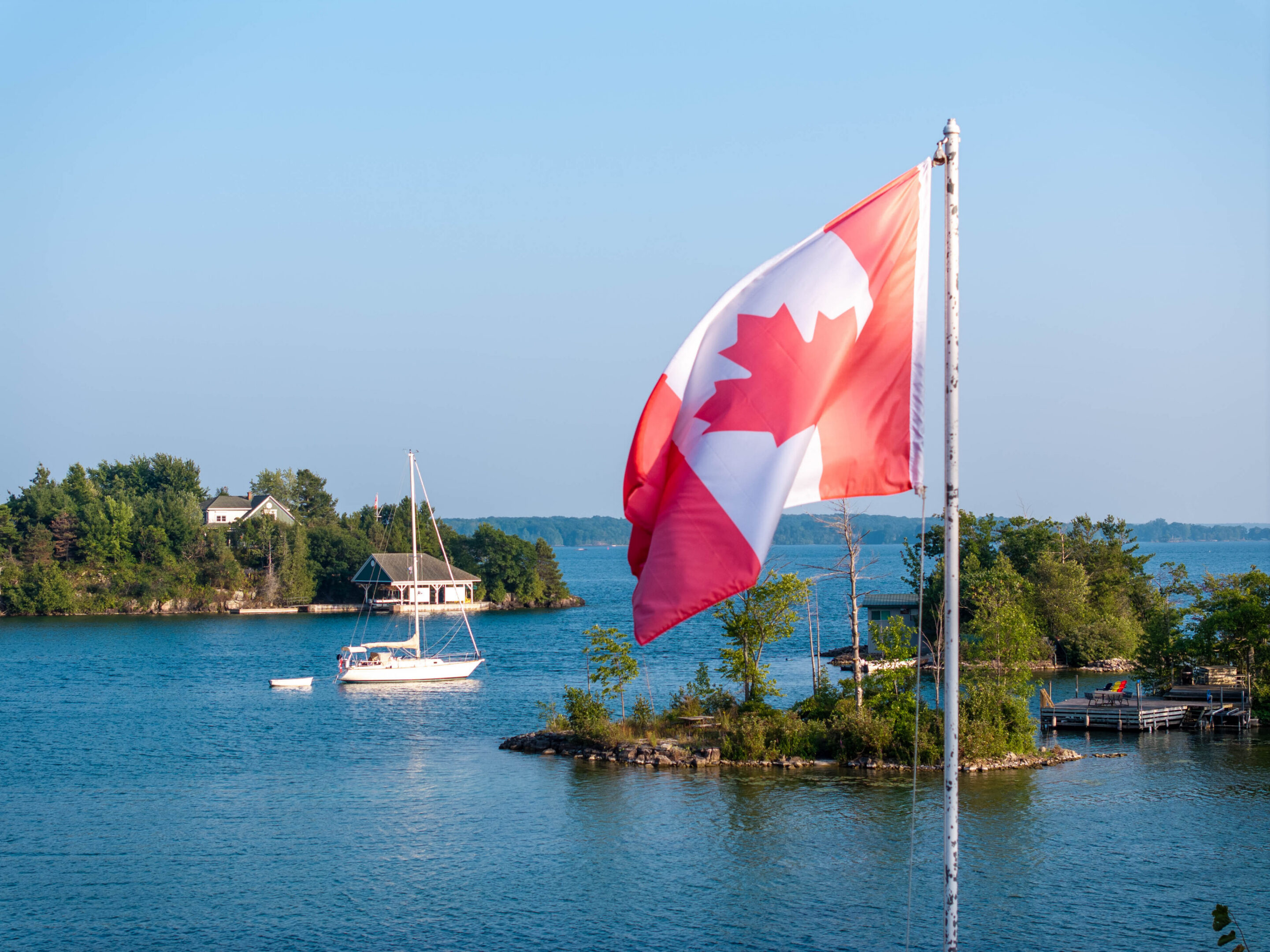 A scenic summer view of the Thousand Islands region in Ontario, Canada, featuring a large Canadian flag waving in the foreground over blue lake water. Small islands with lush green trees, a sailboat, and waterfront cottages create a peaceful and iconic Canadian landscape. This image represents national pride, nature, travel, tourism, and outdoor recreation in Canada